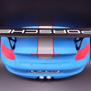 K&ouml;nnte beinhalten: Ein blaues Spielzeugauto mit einem schwarzen Spoiler mit dem wei&szlig;en Schriftzug "PORSCHE". Das Auto hat rote und graue Streifen und Akzente. Das Heck des Autos ist sichtbar, einschlie&szlig;lich Auspuffrohren und Nummernschild.