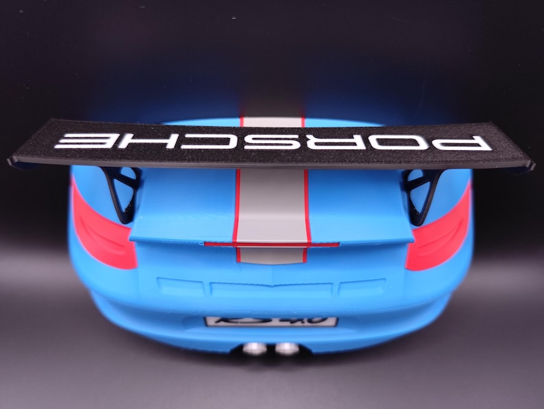 K&ouml;nnte beinhalten: Ein blaues Spielzeugauto mit einem schwarzen Spoiler mit dem wei&szlig;en Schriftzug "PORSCHE". Das Auto hat rote und graue Streifen und Akzente. Das Heck des Autos ist sichtbar, einschlie&szlig;lich Auspuffrohren und Nummernschild.
