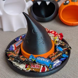 May include: A black witch's hat-shaped candy dish, decorated with a crescent moon and stars, filled with wrapped sweets. The hat sits on an orange base and a black tray. Other Halloween-themed dishes include a ghost, bat, and pumpkin.