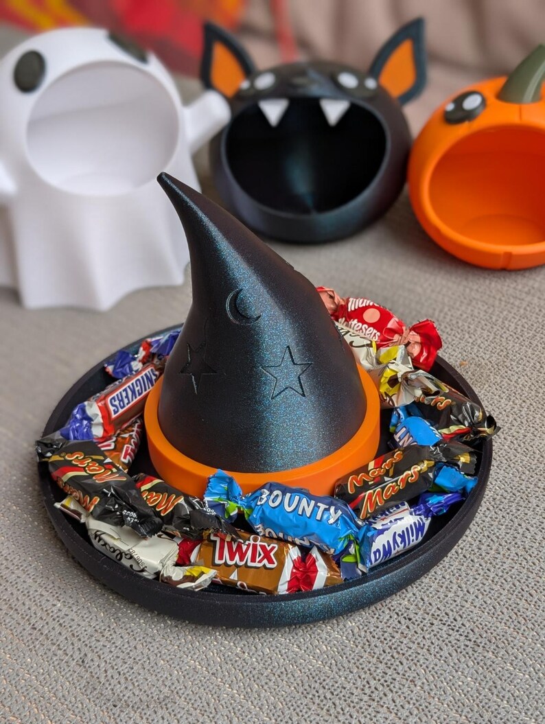 May include: A black witch's hat-shaped candy dish, decorated with a crescent moon and stars, filled with wrapped sweets. The hat sits on an orange base and a black tray. Other Halloween-themed dishes include a ghost, bat, and pumpkin.