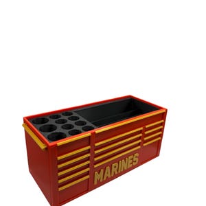 May include: A red and gold tool organizer with the word "MARINES" in gold lettering. The organizer has multiple compartments, including a section with circular holders and a larger open area. The design features horizontal gold stripes.