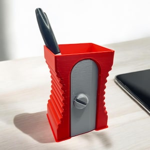 Könnte beinhalten: Ein roter Bleistiftspitzer mit einer grauen Schleifklinge und einem schwarzen Stift im Inneren. Der Spitzer steht auf einer hellen Holzoberfläche.