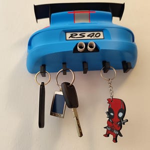 K&ouml;nnte beinhalten: Ein blauer, auto-f&ouml;rmiger Schl&uuml;sselhalter mit dem Text "RS 4.0" und zwei Auspuffrohren. Der Schl&uuml;sselhalter hat vier schwarze Haken mit Schl&uuml;sseln und Schl&uuml;sselanh&auml;ngern. Ein Schl&uuml;sselanh&auml;nger zeigt ein Deadpool-Design.