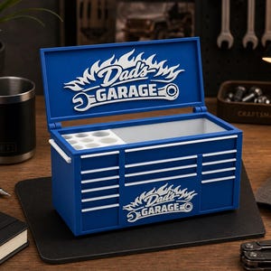 May include: A blue toolbox with white accents and the text "Dad's Garage" in a flame design. The toolbox is open, revealing a white interior with small compartments. A black Yeti tumbler and a notebook are on the table.