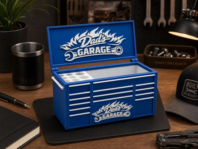 May include: A blue toolbox with white accents and the text "Dad's Garage" in a flame design. The toolbox is open, revealing a white interior with small compartments. A black Yeti tumbler and a notebook are on the table.