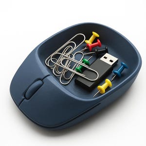 Op de afbeelding: Een marineblauwe bureau-organizer in de vorm van een computermuis, gevuld met zilveren paperclips, een zwarte USB-stick en kleurrijke punaises in rood, geel, groen en blauw. De organizer staat op een witte achtergrond.