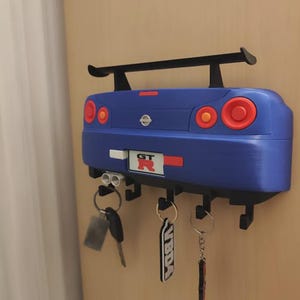 May include: A blue car-shaped key holder with a black spoiler and red and orange taillights. The key holder has several hooks for keys and a "GT R" emblem. Keys are hanging from the hooks.