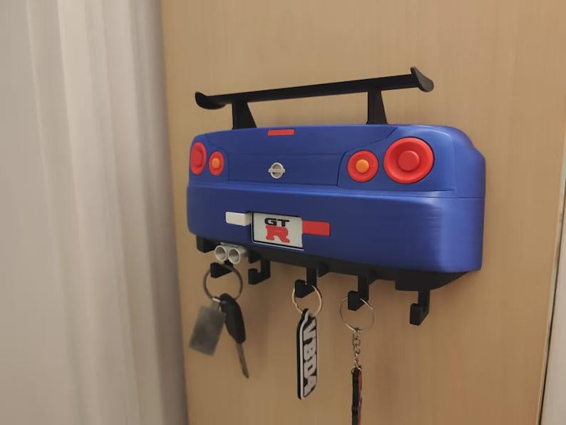 May include: A blue car-shaped key holder with a black spoiler and red and orange taillights. The key holder has several hooks for keys and a "GT R" emblem. Keys are hanging from the hooks.