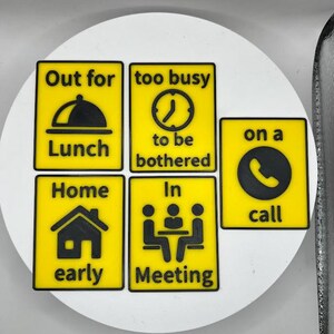 May include: A set of yellow and black signs with various icons and text. The signs read "Out for Lunch", "too busy to be bothered", "on a call", "Home early", and "In Meeting". The signs are arranged on a white surface.