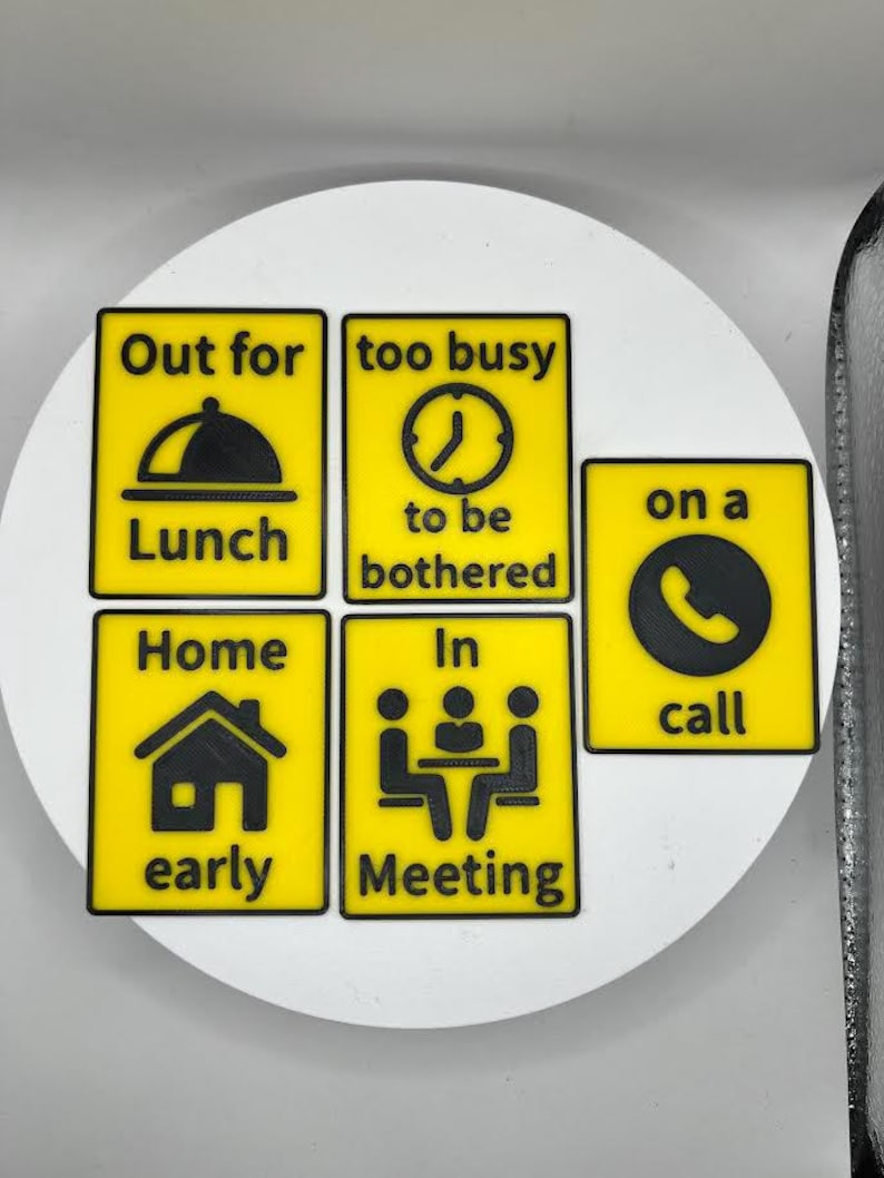 May include: A set of yellow and black signs with various icons and text. The signs read "Out for Lunch", "too busy to be bothered", "on a call", "Home early", and "In Meeting". The signs are arranged on a white surface.