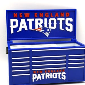 May include: A blue and white New England Patriots toolbox with the team's logo and name prominently displayed. The toolbox has a hinged lid and a series of horizontal lines on the front, with the team name repeated at the bottom.