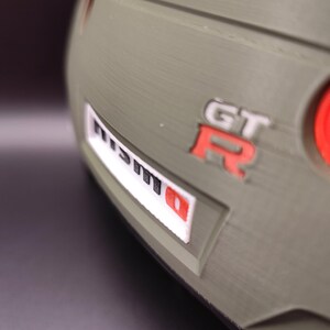 Może przedstawiać: Zbliżenie fragmentu samochodu, z logo "NISMO" w kolorze białym i czerwonym oraz emblematem "GT-R" w kolorze czerwonym. Karoseria samochodu jest w kolorze matowej oliwkowej zieleni.