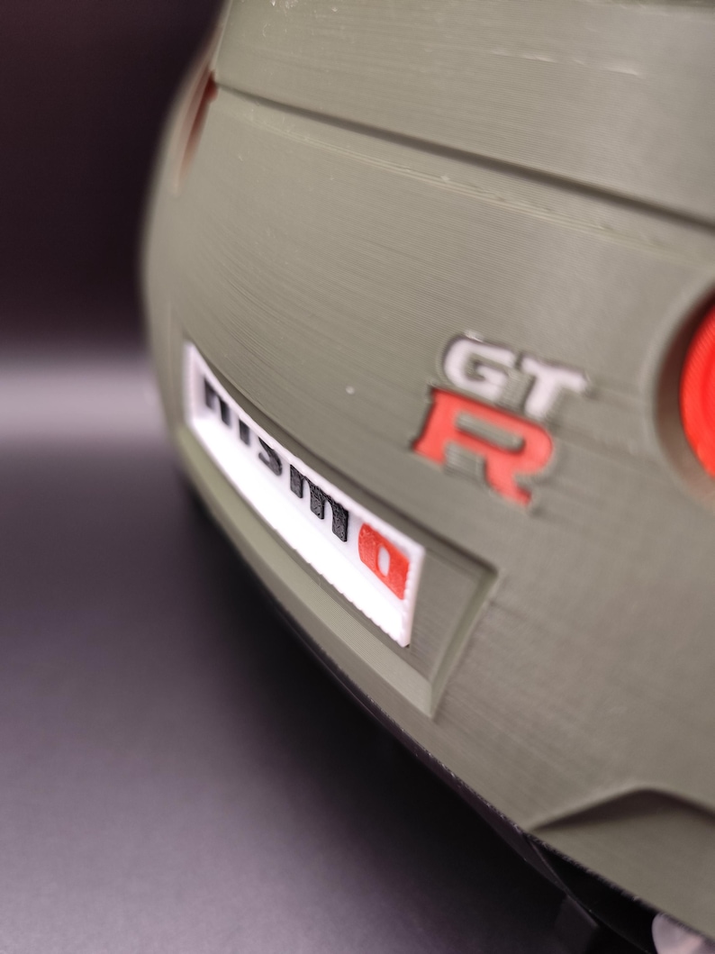 Może przedstawiać: Zbliżenie fragmentu samochodu, z logo "NISMO" w kolorze białym i czerwonym oraz emblematem "GT-R" w kolorze czerwonym. Karoseria samochodu jest w kolorze matowej oliwkowej zieleni.