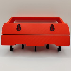May include: A bright orange wall-mounted shelf with a rectangular frame and a lower section with four black hooks. The shelf has two black keyhole-shaped hanging mounts on the back.