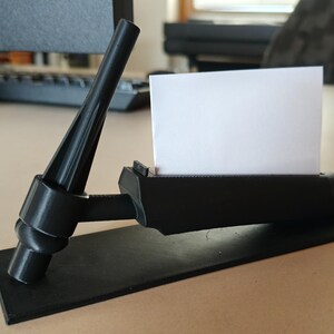 May include: A black business card holder with a unique, angular design. The holder features a rectangular base, a cylindrical section to hold cards, and a support structure. A white business card is inserted in the holder.