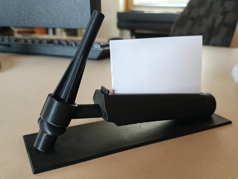 May include: A black business card holder with a unique, angular design. The holder features a rectangular base, a cylindrical section to hold cards, and a support structure. A white business card is inserted in the holder.