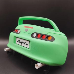 May include: A green model car rear end with the word "Supra" on the license plate. The model has a spoiler, tail lights, and exhaust pipes. The car is a vibrant green color.