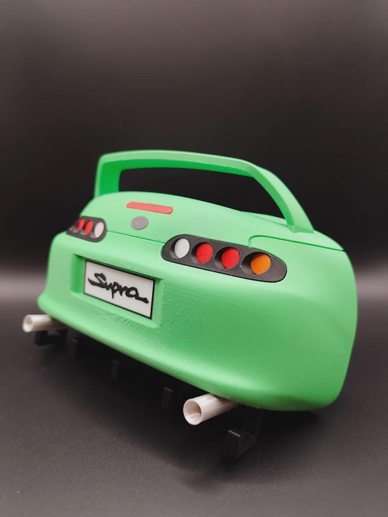 May include: A green model car rear end with the word "Supra" on the license plate. The model has a spoiler, tail lights, and exhaust pipes. The car is a vibrant green color.