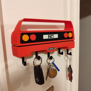 May include: Red key holder shaped like a car, with the text "F40" on it. The key holder has four black hooks and is mounted on a white wall. Several keys hang from the hooks, including a car key and a keychain.