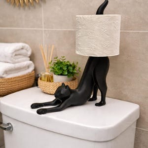 Pode incluir: Um porta-papel higi&ecirc;nico em forma de gato preto. O gato est&aacute; em uma pose de alongamento, com as patas dianteiras estendidas e as pernas traseiras sustentando um rolo de papel higi&ecirc;nico branco. O suporte est&aacute; em um vaso sanit&aacute;rio branco.