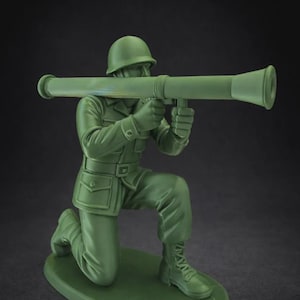 May include: A green plastic toy soldier in a kneeling position, holding a green bazooka. The soldier wears a helmet, uniform, and boots. The toy is on a green base, against a dark background.
