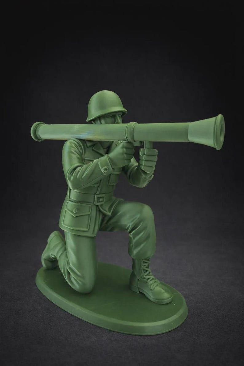 May include: A green plastic toy soldier in a kneeling position, holding a green bazooka. The soldier wears a helmet, uniform, and boots. The toy is on a green base, against a dark background.