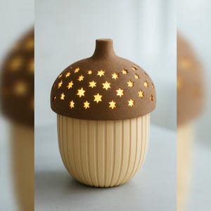 Peut inclure: Lampe décorative en forme de gland, avec un dessus marron et une base crème. Le dessus marron est percé d'étoiles qui diffusent une lumière chaleureuse. La base présente des rainures verticales.