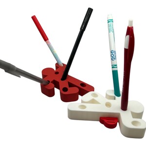 May include: Two red and white plastic pencil holders shaped like stick figures. The holders have holes for pens and pencils. There are four pens and pencils in the holders, including a black pen, a red pen, a blue pen, and a white marker.