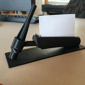 May include: A black 3D-printed business card holder. The holder has a rectangular base, a curved section to hold cards, and a support structure. A white business card is in the holder.