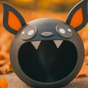 May include: A dark gray, spherical bat-shaped container with large ears, orange inner ears, and white fangs. The container has large, round eyes with white pupils and a small nose. The background is blurred with fall leaves.