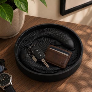 Puede incluir: Una bandeja decorativa redonda y negra con un dise&ntilde;o de carpa koi tallada. La bandeja contiene una cartera de cuero marr&oacute;n, llaves de coche y un llavero. Un reloj de pulsera y gafas de sol son visibles.