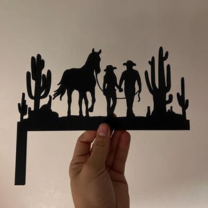 May include: Black metal silhouette of a couple on horseback with cacti in the background. The silhouette is mounted on a metal bracket.