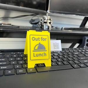 May include: A bright yellow, two-sided sign that reads "Out for Lunch" with a graphic of a covered plate. The sign is placed on a black computer keyboard.