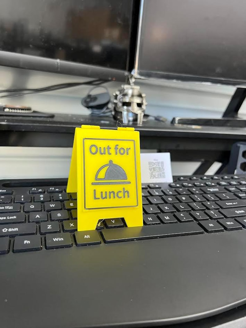 May include: A bright yellow, two-sided sign that reads "Out for Lunch" with a graphic of a covered plate. The sign is placed on a black computer keyboard.