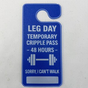 K&ouml;nnte beinhalten: Blauer T&uuml;rh&auml;nger mit dem Text "LEG DAY TEMPORARY CRIPPLE PASS - 48 HOURS -" und einer Hantelgrafik. Auf dem unteren Teil des Anh&auml;ngers steht "SORRY, I CAN'T WALK."