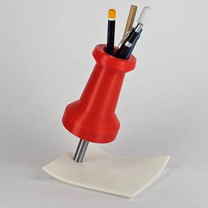 May include: A red pushpin-shaped desk organizer holding pencils and pens. The organizer is 3D-printed and sits on a white, curved base. The pencils and pens are black, yellow, and silver.