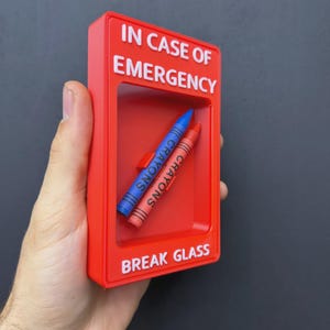 May include: A rectangular red plastic emergency box with white text that reads IN CASE OF EMERGENCY and BREAK GLASS. Inside the recessed center, two wax crayons, one blue and one orange, are positioned diagonally against the red back panel.