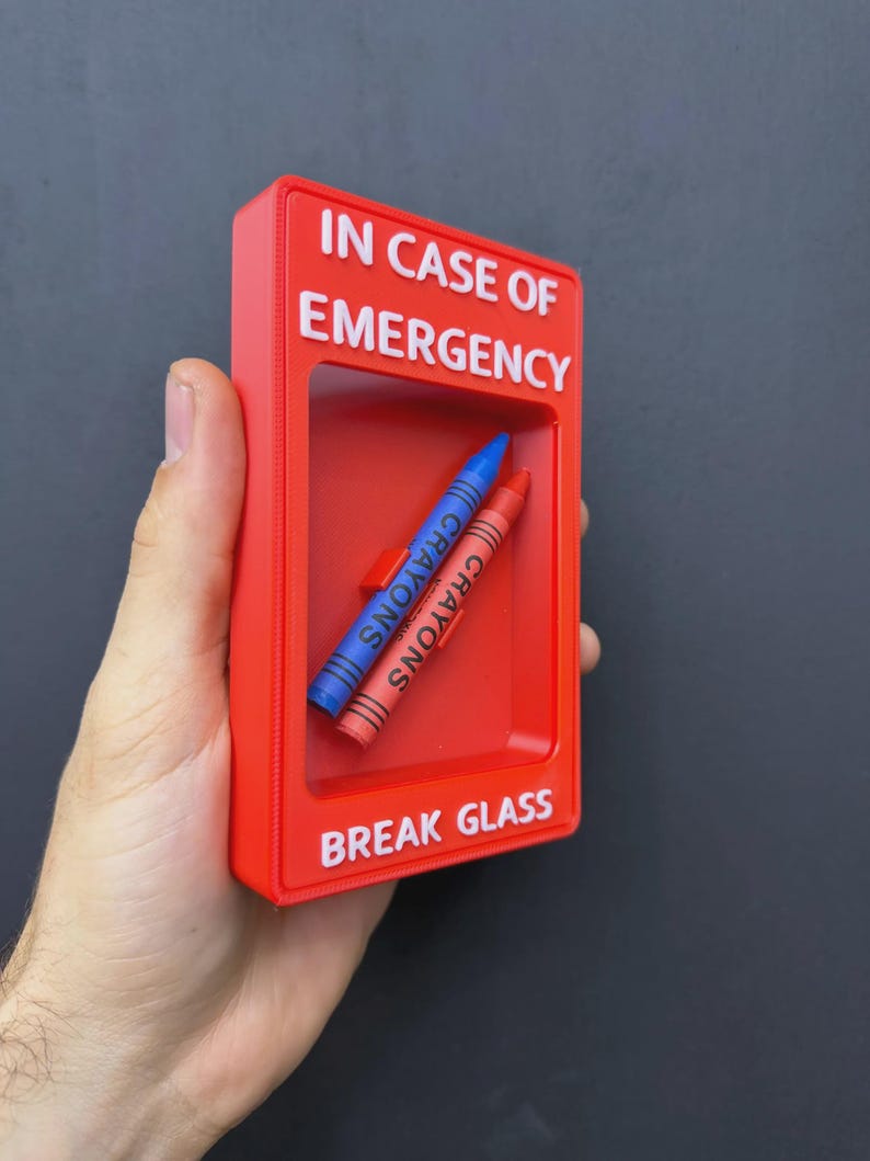 May include: A rectangular red plastic emergency box with white text that reads IN CASE OF EMERGENCY and BREAK GLASS. Inside the recessed center, two wax crayons, one blue and one orange, are positioned diagonally against the red back panel.