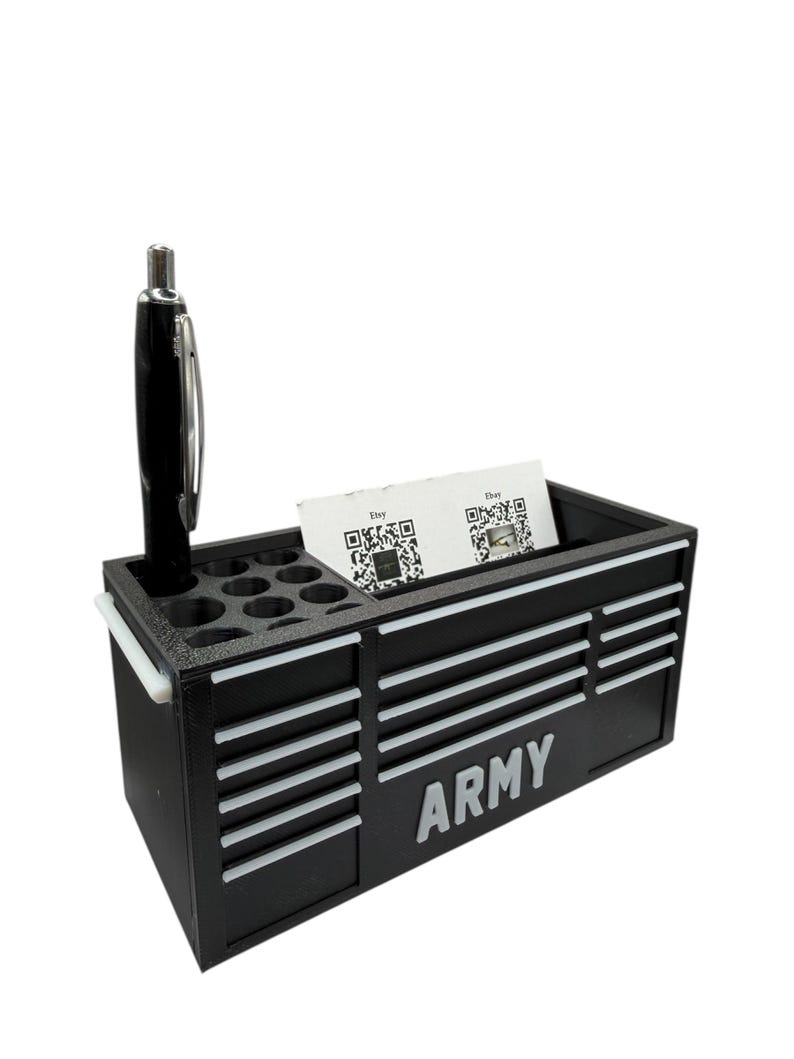 May include: Black desk organizer shaped like a toolbox, with white accents and the word "ARMY" on the front. It has compartments for pens, business cards, and a pen. A black and silver pen is in the pen holder.