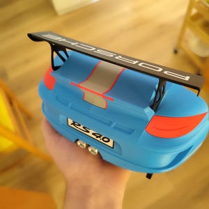 K&ouml;nnte beinhalten: Ein blaues Spielzeugauto mit einem schwarzen Spoiler mit der Aufschrift "PORSCHE". Das Auto hat rote und graue Akzente, und das Nummernschild lautet "RS 4.0". Das Spielzeug wird in einer Hand gehalten.
