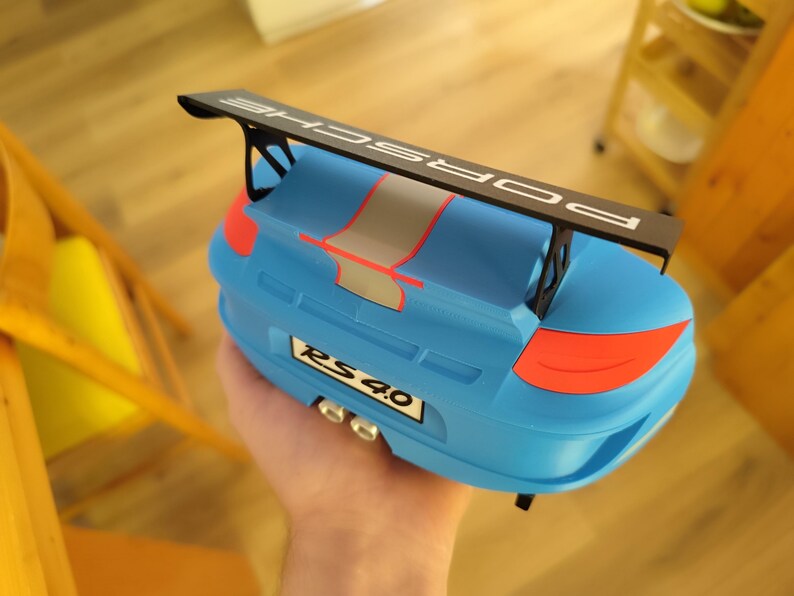 K&ouml;nnte beinhalten: Ein blaues Spielzeugauto mit einem schwarzen Spoiler mit der Aufschrift "PORSCHE". Das Auto hat rote und graue Akzente, und das Nummernschild lautet "RS 4.0". Das Spielzeug wird in einer Hand gehalten.