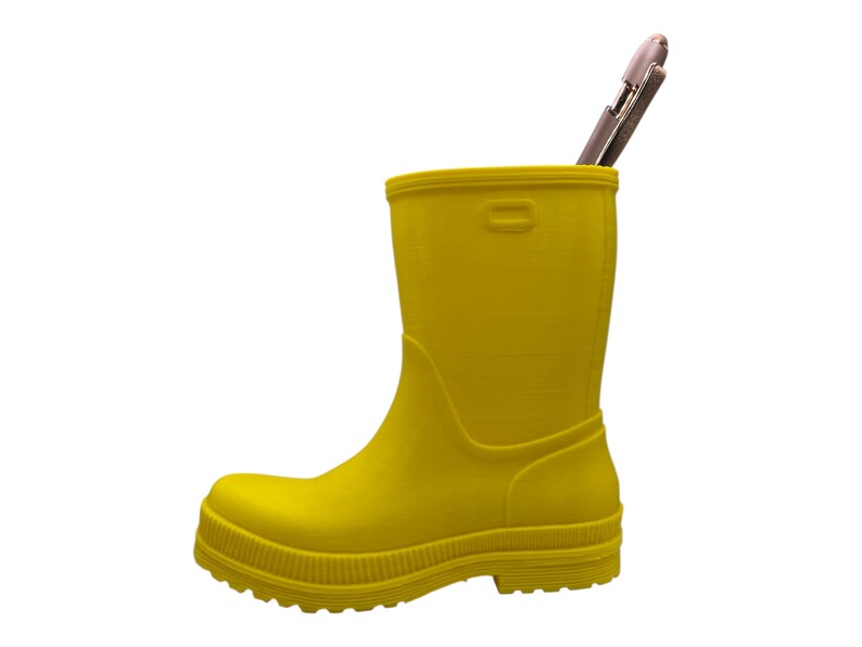 May include: A bright yellow rubber boot used as a pen holder. A silver pen is inside the boot. The boot has a textured surface and a ridged sole. The boot is approximately 8 inches tall.
