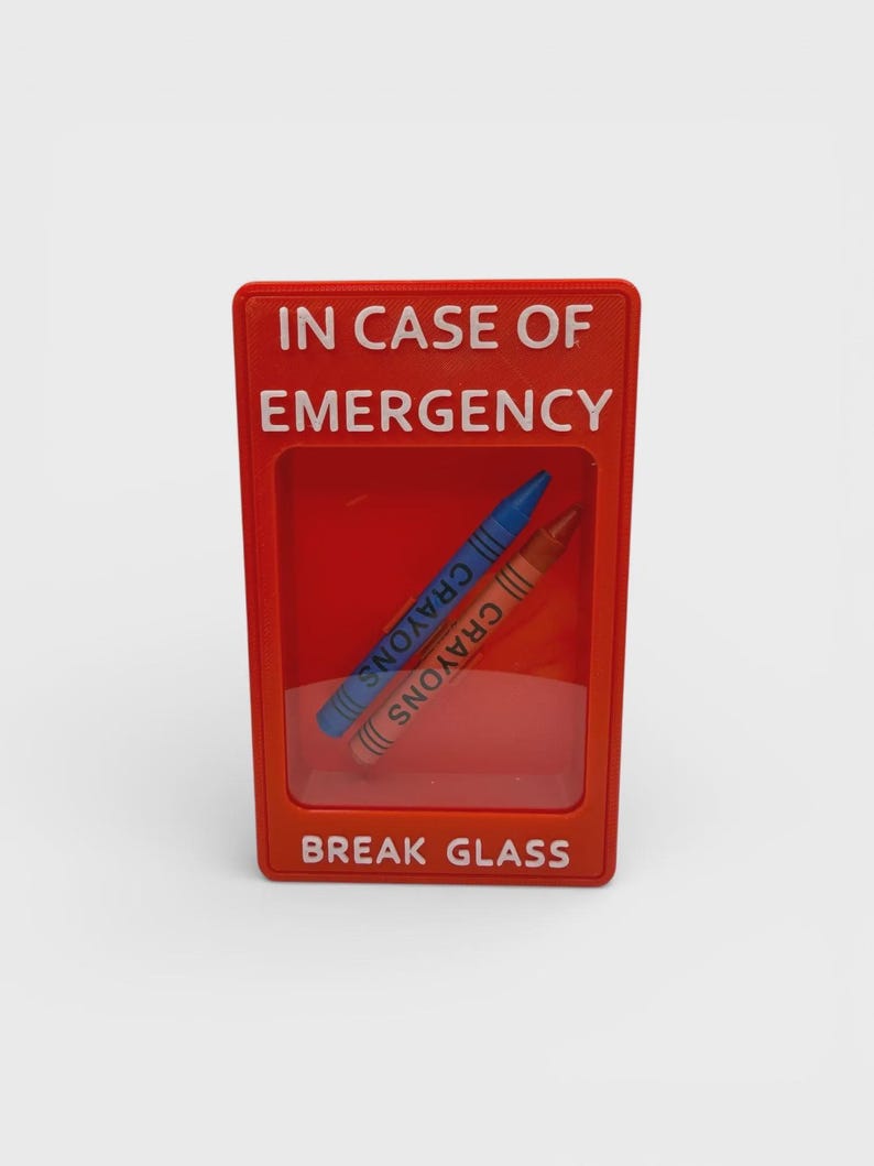 May include: A rectangular red plastic display case with a clear front window. The text IN CASE OF EMERGENCY is printed at the top and BREAK GLASS at the bottom in white. Inside, two wax crayons, one blue and one brown, are positioned diagonally.