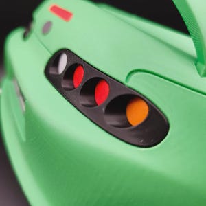 May include: Close-up of a green toy car with a black headlight housing. The car features red, white, and orange lights. A green spoiler and handle are visible. The car's body has a textured surface.