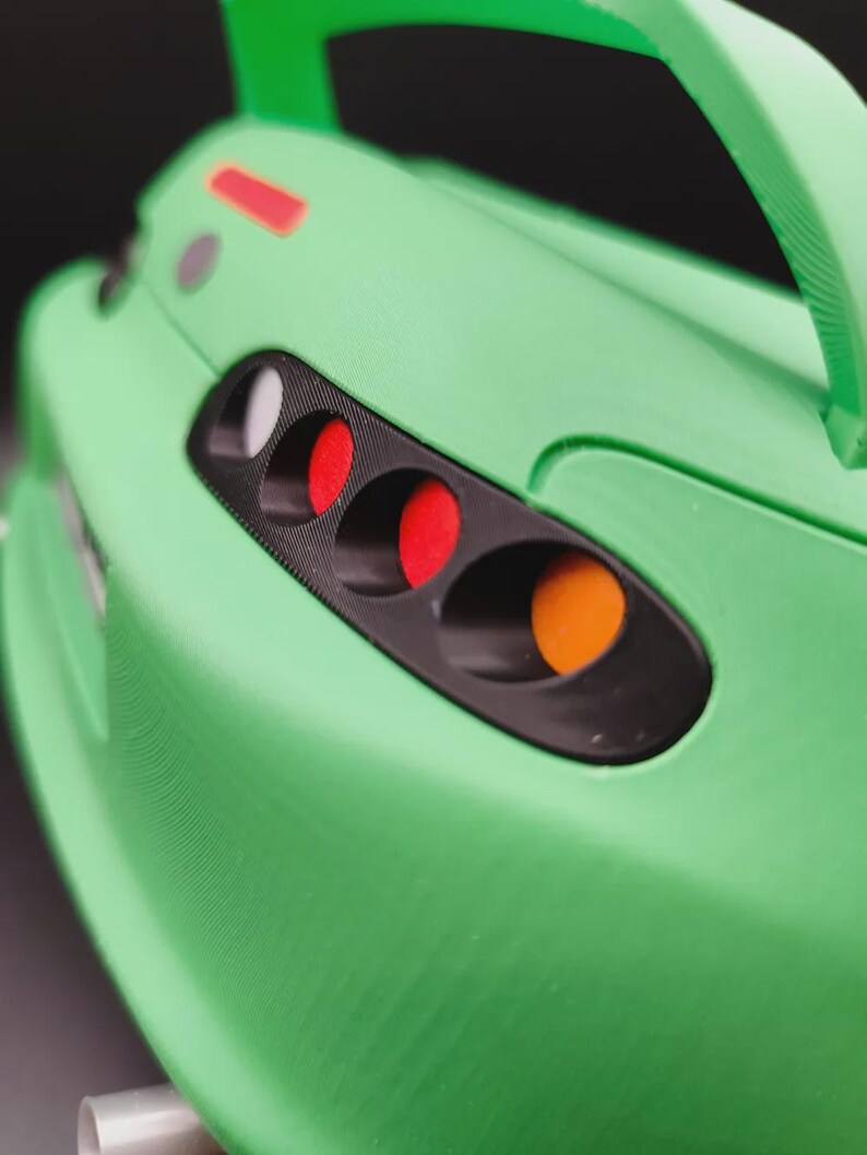 May include: Close-up of a green toy car with a black headlight housing. The car features red, white, and orange lights. A green spoiler and handle are visible. The car's body has a textured surface.
