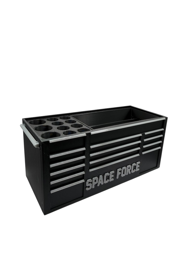 May include: Black tool chest with silver accents and the words "SPACE FORCE" in silver. The chest has multiple drawers and compartments for storage, including a section with circular holders. A handle is on the side.