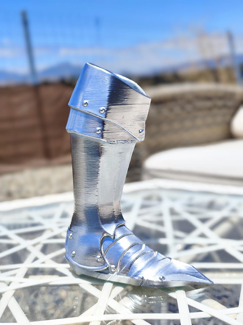 May include: A polished, silver-coloured metal leg armour piece. The armour features a detailed design with multiple plates and rivets, and a pointed toe. The upper part of the armour has a flared design. The piece is displayed on a glass table with a blurred outdoor background.
