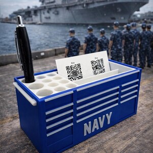 May include: A blue desk organizer with white accents and the word "NAVY" in white. The organizer holds a black pen and business cards with QR codes. A military ship and uniformed personnel are in the background.