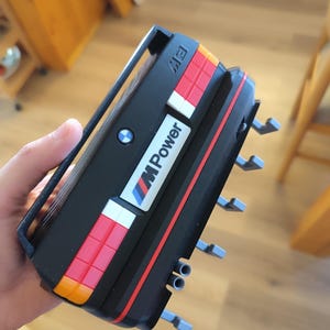 May include: A black and red model car with the text "M Power" and the BMW logo. The model has orange, red, white, and blue accents. The model has a handle on top and several gray protrusions on the side.