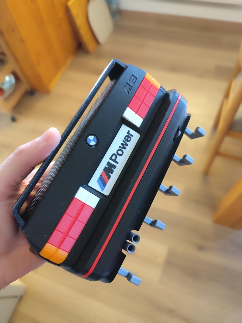 May include: A black and red model car with the text "M Power" and the BMW logo. The model has orange, red, white, and blue accents. The model has a handle on top and several gray protrusions on the side.